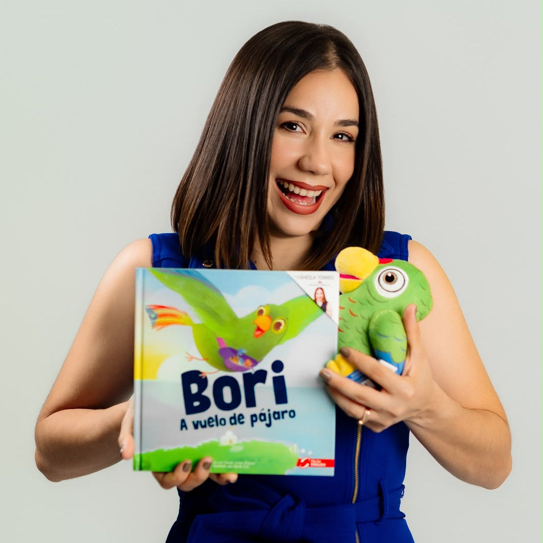 Fotografía de la autora Sheila Torres sonriendo felizmente mientras sostiene el libro infantil bilingüe "Bori: A Vuelo de Pájaro" en una mano y un peluche verde de la cotorra protagonista en la otra.