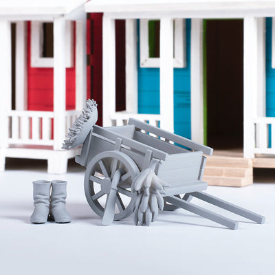 Fotografía de estudio de piezas del set Mini-Accesorios del Campo sin pintar, mostrando la carreta con racimo de plátanos y las botas, con casitas de campo de fondo.