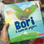 Fotografía compartida por un cliente sosteniendo en sus manos el libro "Bori: A Vuelo de Pájaro", mostrando el tamaño real del producto y los colores vibrantes de su portada.
