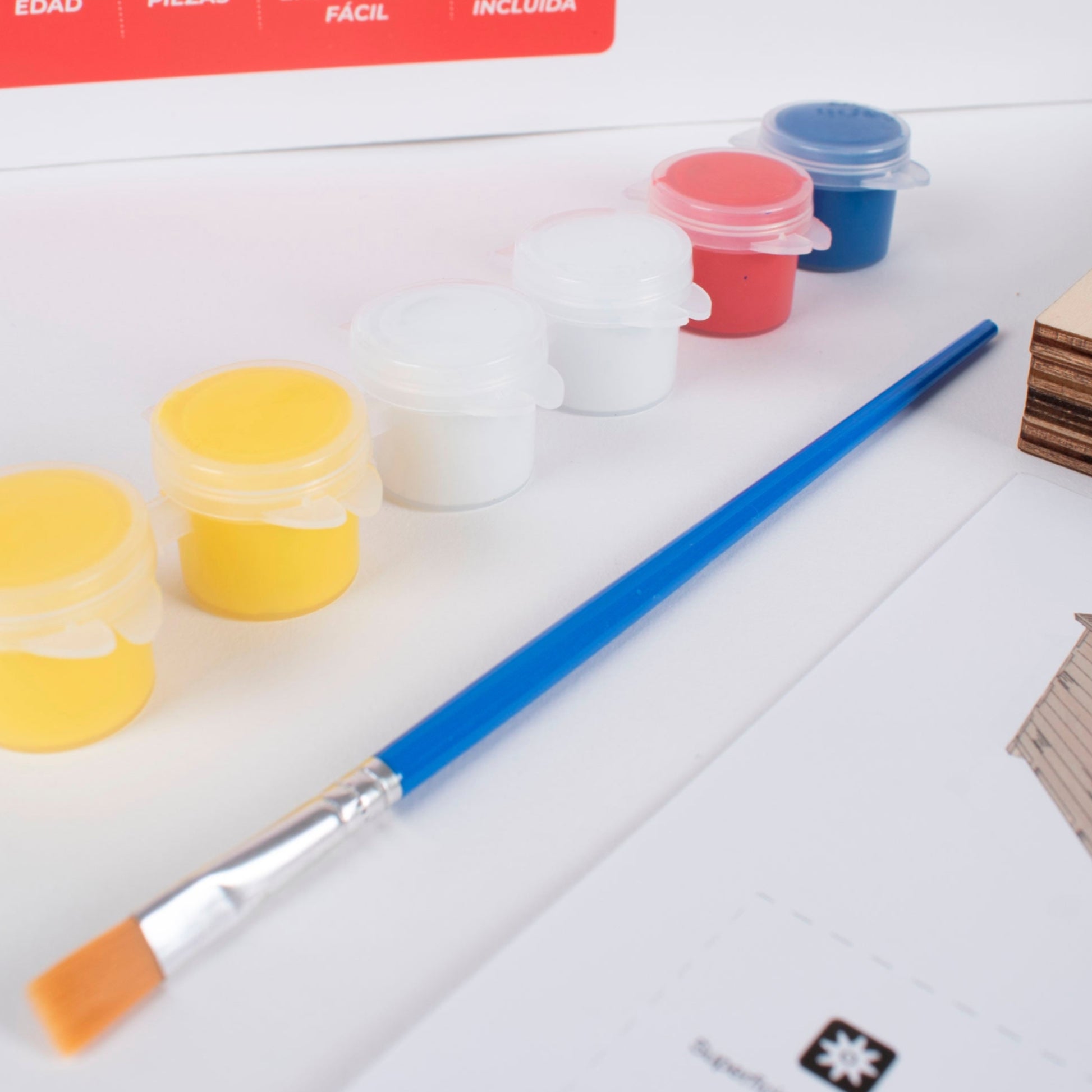 Fotografía en primer plano de los materiales del kit Casita de Campo Modelo Alegría, destacando el set de pinturas, una brocha, los paneles de madera precortados, parte de las instrucciones y el empaque.