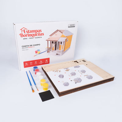 Fotografía de los componentes del kit DIY Casita de Campo Modelo Alegría junto a su empaque, mostrando los paneles de madera procesada, el manual de instrucciones, el set de pinturas, brochas y una lija.