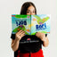 Fotografía divertida de la autora asomando sus ojos por encima del libro "Bori: A Vuelo de Pájaro" abierto de par en par, mostrando el formato bilingüe reversible de las portadas mientras viste una camiseta de Dialecto Boricua.