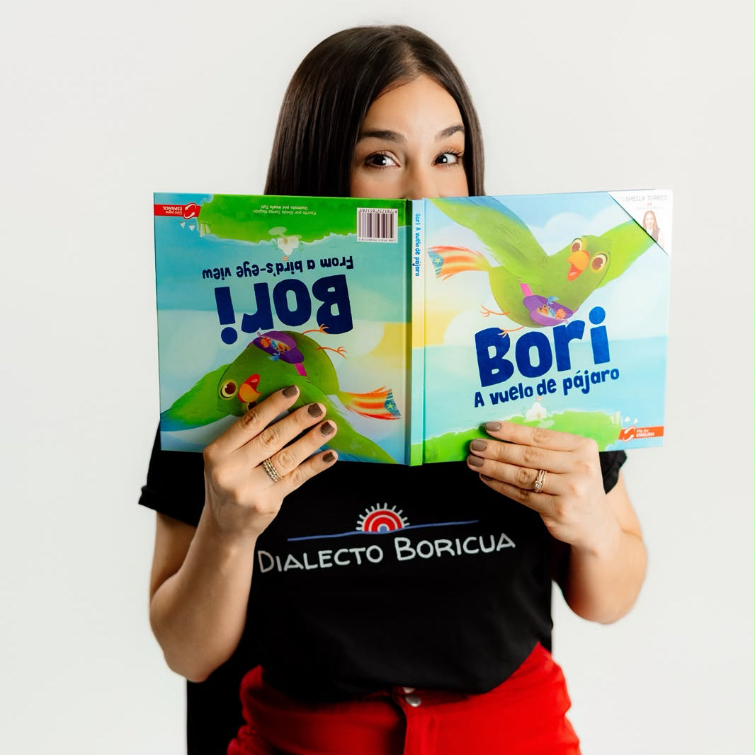 Fotografía divertida de la autora asomando sus ojos por encima del libro "Bori: A Vuelo de Pájaro" abierto de par en par, mostrando el formato bilingüe reversible de las portadas mientras viste una camiseta de Dialecto Boricua.