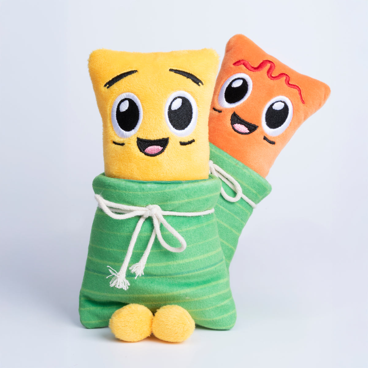 Fotografía de estudio de los peluches Yuca y Masa abrazados, envueltos en sus hojas verdes de tela, mostrando sus caritas bordadas sin ketchup.