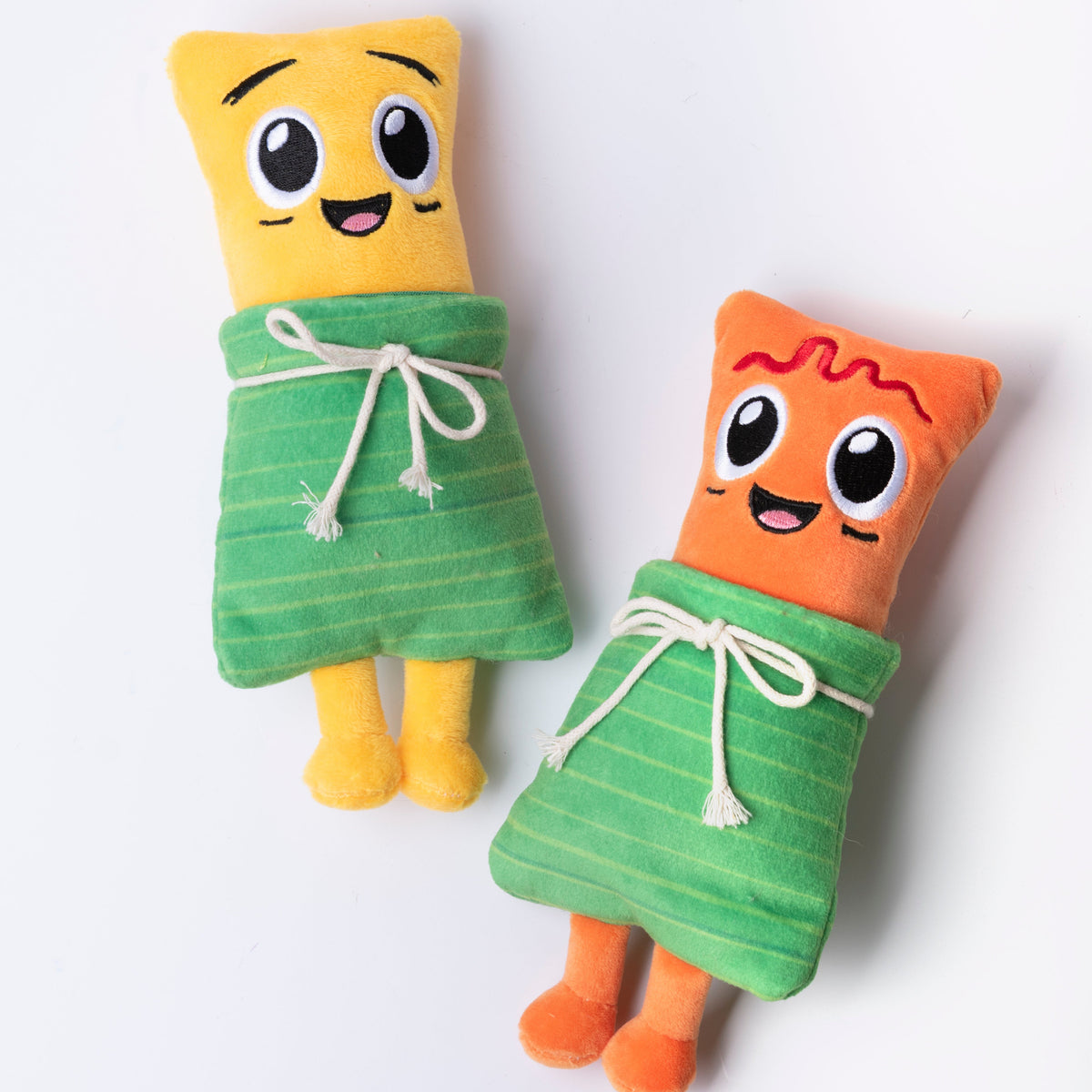 Fotografía de estudio de los peluches Yuca y Masa acostados, mostrando su diseño completo con piernas y hoja verde de tela envolvente.