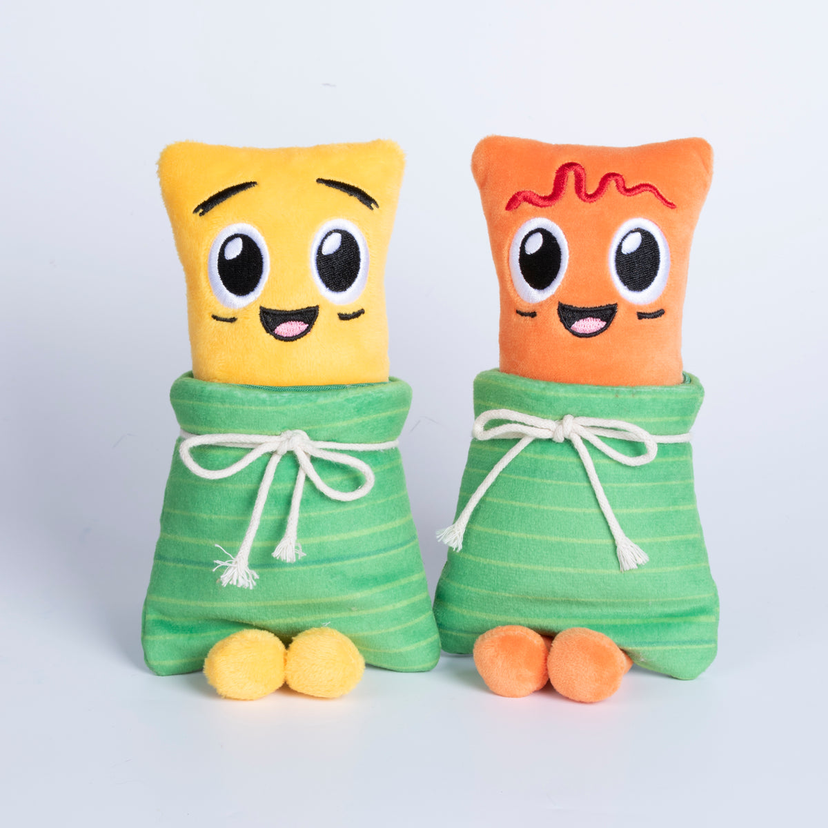 Fotografía de estudio de los peluches Yuca (amarillo) y Masa (anaranjado) sentados lado a lado, envueltos en su hoja verde de tela y mostrando el lado sin ketchup.