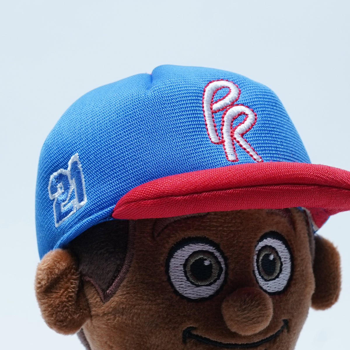 Fotografía de estudio de un acercamiento a la gorra azul con visera roja del peluche de Clemente, con las letras PR y el número 21 bordados.
